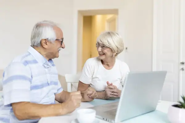 Couple âgé souriant devant un ordinateur, discutant de leur compte bancaire sous curatelle renforcée.
