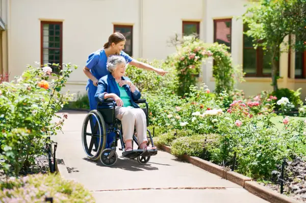 Une aide-soignante pousse une dame âgée en fauteuil roulant dans un jardin fleuri. Une scène paisible, loin des préoccupations de placement en EHPAD sans consentement.