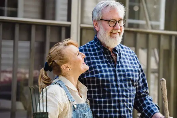 Couple souriant devant une petite maison pour personne âgée. L'homme porte une chemise à carreaux, la femme une salopette en jean.