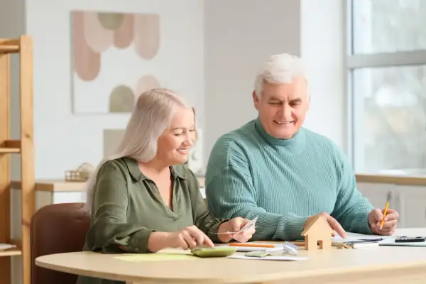 Couple âgé planifiant leur budget pour l'entrée en EHPAD et la résidence principale, avec une calculatrice et une petite maison en bois.
