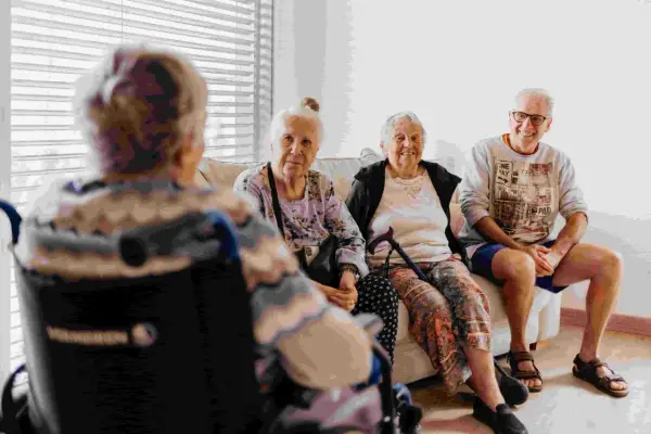 Quatre personnes âgées discutent sur un canapé. L'une est en fauteuil roulant. Le tarif du foyer logement senior est abordé.