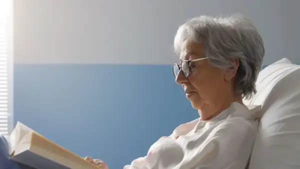 Une femme âgée, en **had et apa**, lit un livre confortablement installée dans un lit.