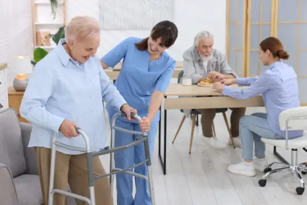 Une aide-soignante accompagne une résidente d'un EHPAD Alzheimer avec un déambulateur. Au fond, d'autres résidents sont à table.