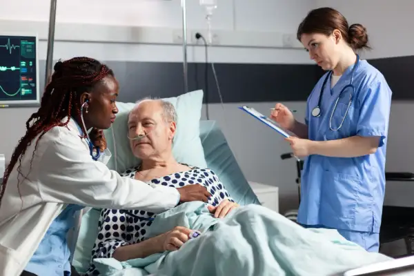 Soins attentifs pour une **hospitalisation à domicile cancer phase terminale**. Une infirmière écoute le cœur d'un patient âgé, une autre note des informations.
