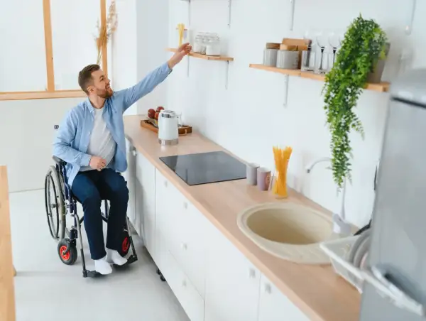 Un homme en fauteuil roulant attrape un objet sur une étagère dans une cuisine adaptée. Un exemple de priorité handicap logement social.