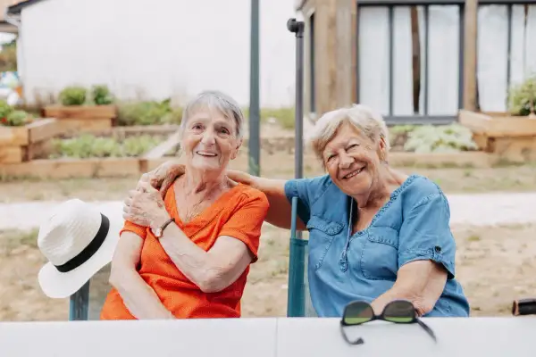 Deux femmes âgées souriantes, profitant de leur logement pour seniors en plein air. Une chapeau de paille est posé sur une chaise.