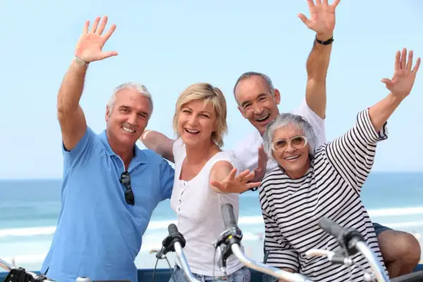 Quatre seniors joyeux agitent les mains, profitant d'une belle journée au bord de la mer, peut-être après une balade à vélo dans ce village senior bord de mer.