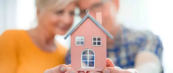 Un couple regarde une maison miniature rose, symbolisant leur recherche d'un logement senior pas cher pour leur avenir.